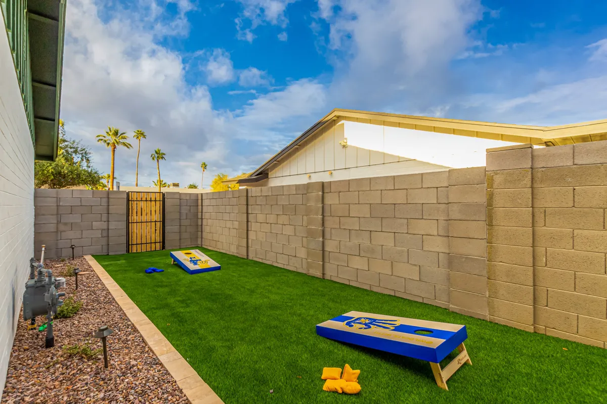 A backyard scene with green artificial grass, cornhole game boards, tall palm trees, and a fenced area. The setting is designed for outdoor games, sunny relaxation, and casual gatherings, radiating a playful and relaxed mood ideal for groups.