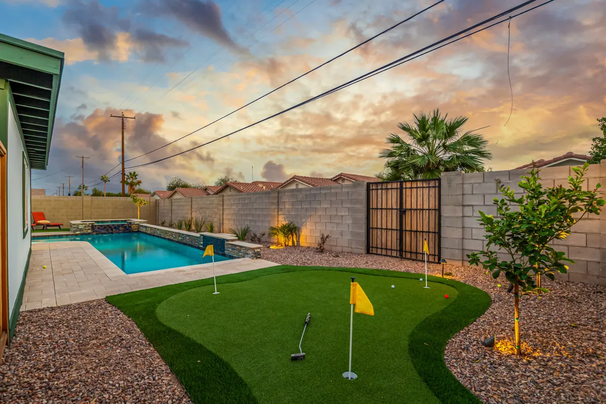 A vibrant exterior shot featuring a shimmering pool, putting green, palm trees, and a beautiful sunset sky. The fenced area and open space suggest opportunities for both recreation and informal team bonding, with plenty of room for evening gatherings or friendly competitions.