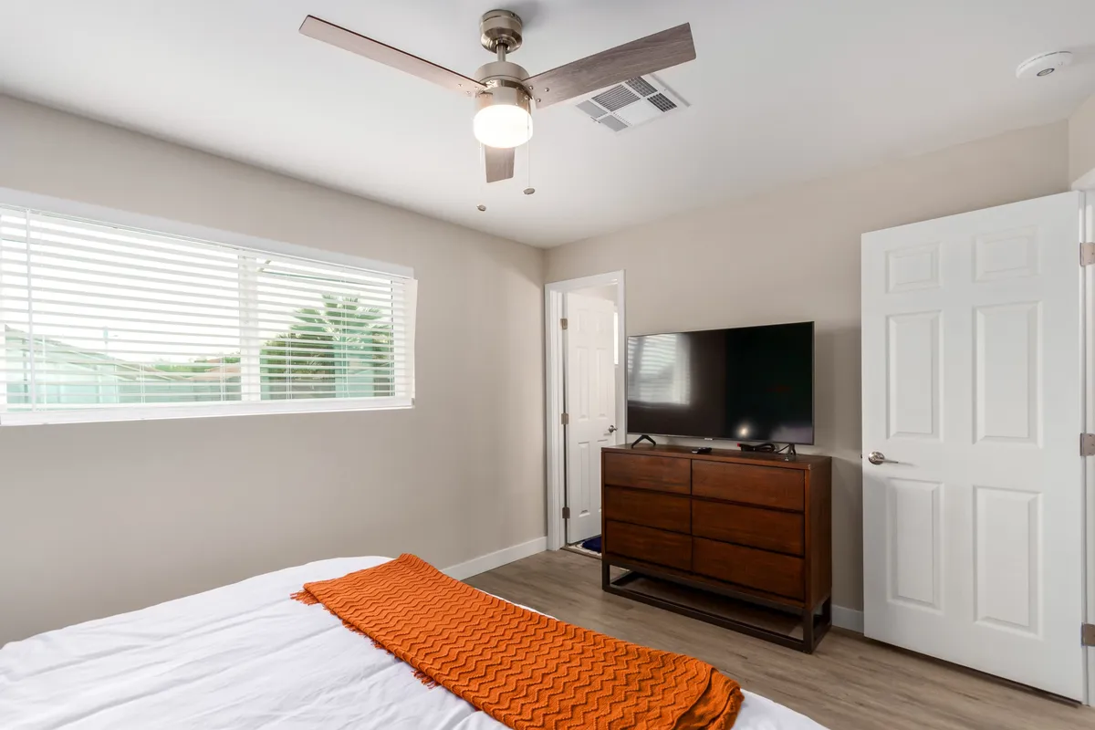 A comfortable bedroom featuring a cozy bed with a vibrant orange throw blanket, a sturdy wooden dresser, and a ceiling fan overhead. The space feels warm and inviting, perfect for unwinding after a day out. Soft lighting and simple decor create an atmosphere conducive to relaxation and restful sleep.