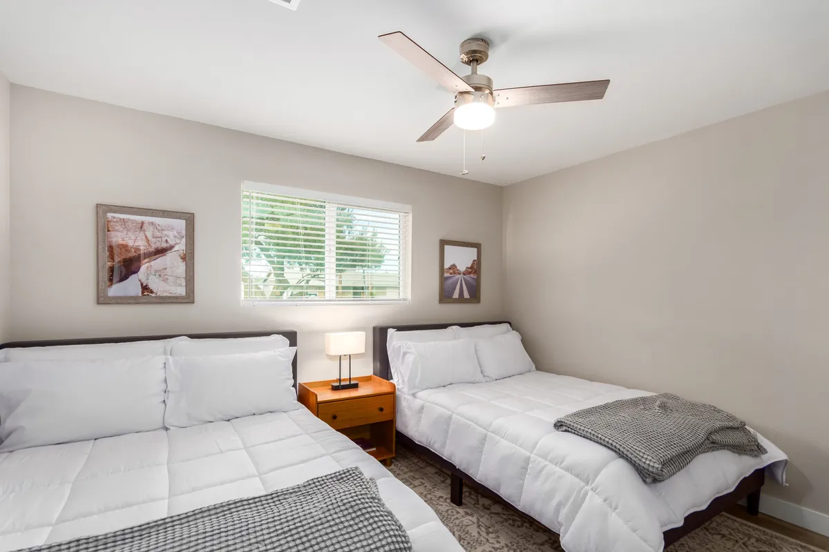 A bright, cheerful bedroom with two beds, a nightstand and lamp between them, a window letting in greenery and natural light, and framed wall art above. The room feels welcoming and ideal for families or friends sharing the space.