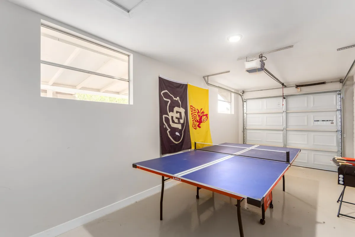 This interior view features a ping pong table positioned near a window, flanked by flags, creating a playful yet relaxed setting. The room is set up for socializing, watching sports, and casual hangouts, spotlighting both recreation and camaraderie.